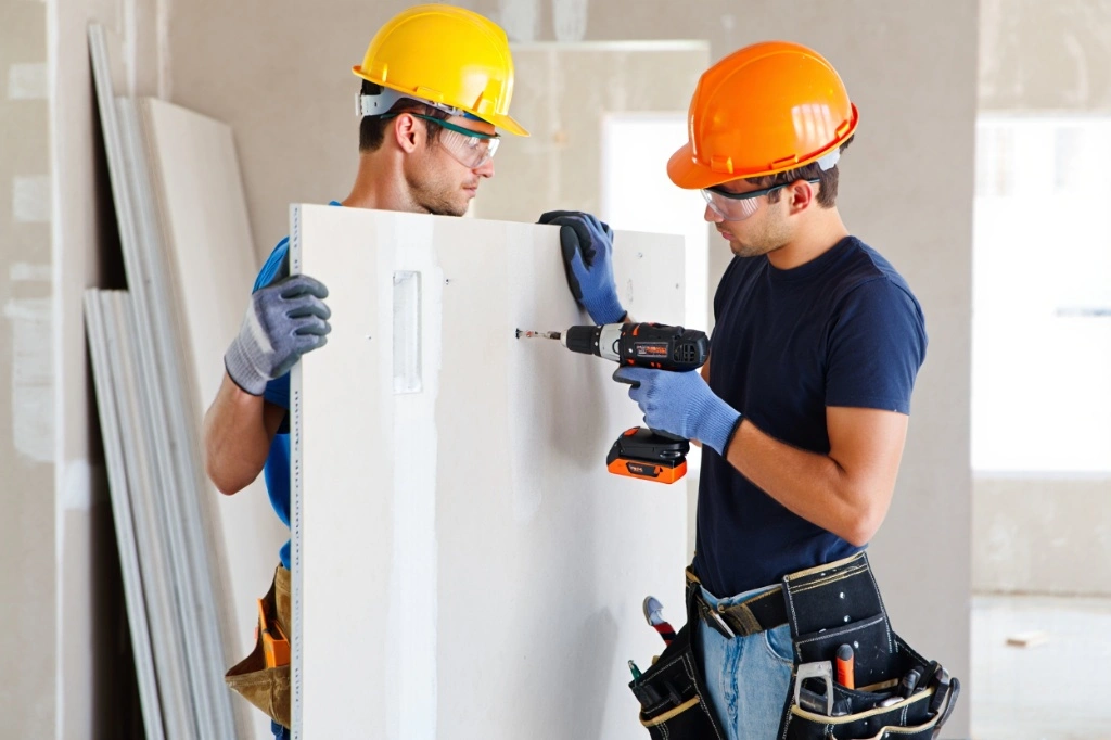  Gemini said Two construction workers wearing hard hats and safety gear installing a drywall access panel using a power drill.